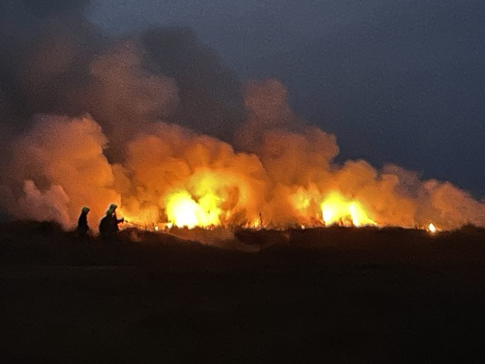 Image of the heath fire on Bryher in full flame at night, with 2 fire fighters uses hoses to extinguish it.